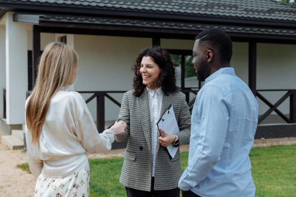 A real estate agent from SMM Realtors, dressed in a checkered blazer, shakes hands with a female client while a male client observes. They are standing outside a modern house with a dark roof and wooden porch, discussing real estate services and FAQs.