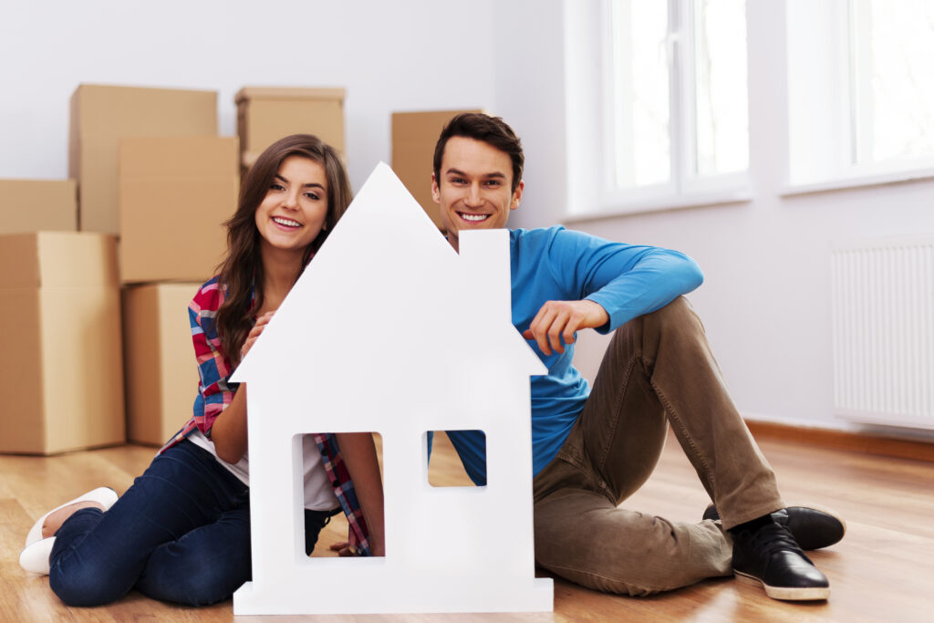 A happy young couple sitting on the floor with a white house-shaped cutout, surrounded by moving boxes in their new home in Siliguri.